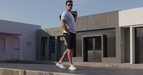 Person in gray t-shirt and shorts walking past light-colored building with 'Cotton' sign. Wearing white sneakers on sunny day.