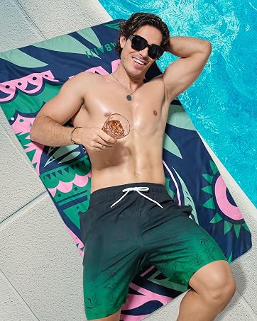 Tropical print swim shorts in dark green with pink accents, shown laid on a patterned beach towel next to a pool.