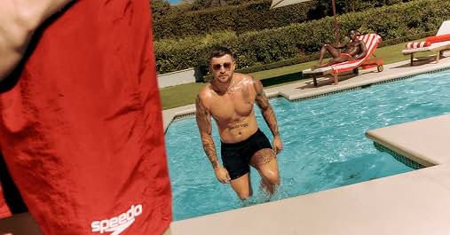 Swimming pool scene with black swim shorts and partial red Speedo garment visible. Pool has lounge chairs along the edge and manicured hedges in background.